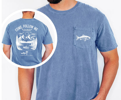 May include: A blue short-sleeved t-shirt with a pocket featuring a white fish graphic. The back of the shirt displays a white graphic with the text "COME, FOLLOW ME" and a mountain and lake scene with a boat, fish, and cross.