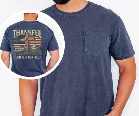 May include: Navy blue short-sleeve t-shirt with a pocket on the chest. The back of the shirt features a graphic with a cross, American flag, mountains, and the text "THANKFUL" and "GOD IS IN CONTROL".